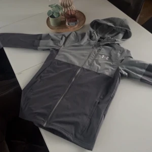 Grå vindjacka från Under Armour - Snygg tvåtonad grå vindjacka från Under Armour med huva och hel dragkedja. Jackan har en diskret logga på bröstet, praktisk bröstficka med dragkedja och sidofickor. Perfekt för dig som gillar sportig stil och vill ha något lätt och funktionellt.
