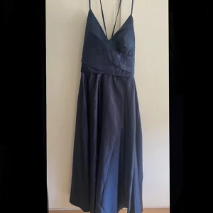 Balklänning - Stilren balklänning i mörkblå/marinblå färg. Tight upptill och löst fallande nedtill med ett snitt vid benet. V-ringad urringning fram och öppen rygg med knytband och diskret dragkedja - ger både stil och komfort. Utan ärmar, med tunna band över axlarna. Material: 100% polyester.  Mått (ungefär storlek M): Totalhöjd (barfota): 157 cm. Nyckelben till golv: 132 cm. Midja: 80 cm. Höfter: 100 cm. Byst: 86 cm.  Öppen för att diskutera pris.  Perfekt för bal, bröllop eller annan formell fest!
