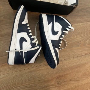 Nike Air Jordan 1 Mid Marinblå/Vit - Säljer ett par Nike Air Jordan 1 Mid i marinblått och vitt. Skorna har klassisk snörning, rund tå och platt sula. Tillverkade i skinn med perforerad tåbox för extra ventilation. Snygg kontrast mellan de vita och marinblå panelerna samt ikonisk Air Jordan-logga på sidan.(PRIS KAN DISKUTERAS)