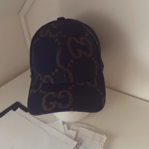 Gucci keps med GG-mönster, marinblå - Snygg marinblå keps från Gucci med klassiskt GG-mönster i brunt över hela kepsen. Justerbar rem baktill med metallspänne. Kepsen har böjd skärm och är tillverkad i jeansmaterial som ger en cool och lyxig vibe.