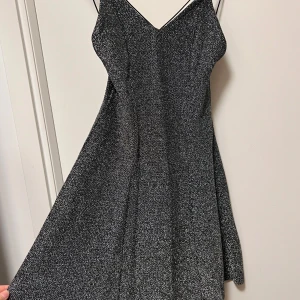 Glittrig svart kortklänning med smala band - Supersnygg svart glittrig klänning med smala axelband och v-ringad front. Klänningen har en lätt utställd form och öppen rygg med korsade band. Perfekt för dig som vill sticka ut med lite extra glitter och glam på nästa event!