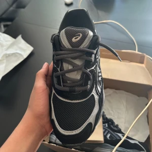 Svarta och grå ASICS sneakers - Säljer ett par ASICS sneakers i svart och grått med mesh och syntetpaneler. Skorna har snörning, rund tå och en rejäl platt sula som ger bra stöd. Snyggt mönstrade detaljer på sidorna och klassisk ASICS-logga på tungan. Perfekt för dig som gillar sportig stil.