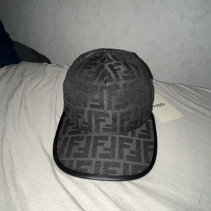 Svart Fendi keps med FF-mönster - Snygg svart keps från Fendi med det klassiska FF-logomönstret över hela kepsen. Kepsen har böjd skärm med svart kant och justerbar rem bak med Fendi-logga. Materialet är bomull och polyester, vilket ger en lyxig och bekväm känsla.