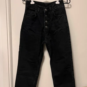 Svarta högmidjade jeans med knappar - Snygga svarta jeans med hög midja och raka ben. Jeansen har fyra synliga silverfärgade knappar framtill och klassiska fem fickor. Perfekta för dig som gillar en clean och stilren look. Materialet är jeans och passformen är normal.