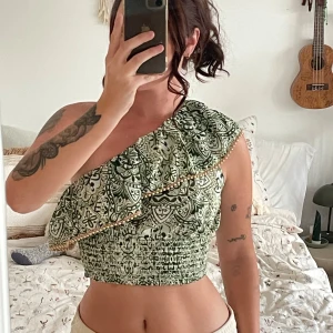 Grön mönstrad oneshoulder topp Primark - Säljer en grön och vit oneshoulder topp från Primark med snyggt mönster och volangkant dekorerad med små pärlor. Toppen är croppad och har smockad midja för en bekväm passform. Ny med prislapp