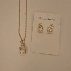 Guldigt halsband och örhängen med pärlor - Snyggt set med halsband och örhängen i guldtonad metall, dekorerade med vita pärlor och små glittriga stenar. Hängena har en elegant, böjd form som ger en lyxig vibe. Perfekt för dig som vill sticka ut med stilrena detaljer och lite extra bling.