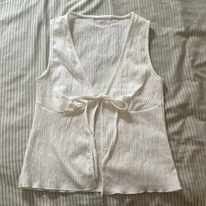 Vit omlottväst med knyt - Säljer en fräsch vit väst i mjukt, strukturerat tyg med omlott-design och knyt framtill. Västen har v-ringad hals och är ärmlös, perfekt att styla över en t-shirt eller klänning. Enkel och clean look som funkar året runt.