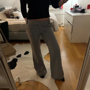 Grå kostymbyxor - Snygga grå kostymbyxor med diskret mönster och två dekorativa knappar bak. Byxorna har låg midja, raka ben med lite bootcut-vibe och hällor för bälte. Materialet känns som en blandning av bomull och syntet, perfekt för en clean och stilren look. Storlek 10 men skulle säga att det är S