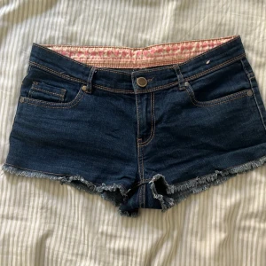 Mörkblå jeansshorts med fransad kant - Snygga mörkblå jeansshorts med fransad kant och klassiska fem fickor. Shortsen har synliga kontrastsömmar och knappstängning fram. Insidan av midjan har ett rosa mönstrat tyg som ger en extra detalj. Perfekta för varma dagar och chill stil.