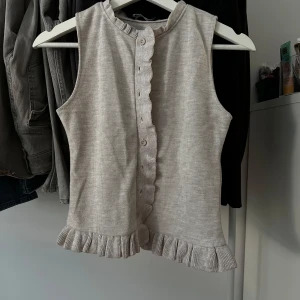 Beige ribbad linnetopp med volang - Supersöt beige ärmlös linnetopp med ribbad struktur, knappar framtill och volangdetaljer längs knappslån och nederkanten. Perfekt för dig som gillar en feminin och chill stil.