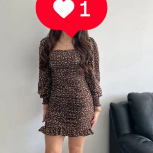 Brun blommig kortklänning med volang - Supersöt brun kortklänning med små vita blommor och volangkant nedtill. Klänningen har långärmade puffärmar och en smickrande, rynkad passform som framhäver figuren. Perfekt för dig som gillar romantiska och trendiga detaljer.