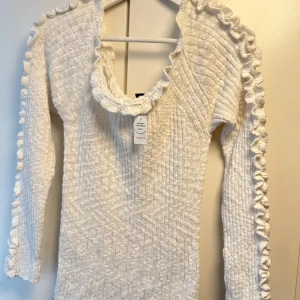 Vit stickad tröja med volang från Lindex - Supersöt vit stickad tröja från Lindex i storlek XL. Tröjan har långärmade ärmar och snygga volangdetaljer längs axlar och halsringning. Ribbad struktur ger en skön känsla och extra stilpoäng. Perfekt för dig som gillar romantiska och trendiga plagg.