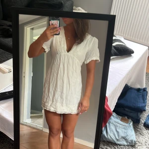 Vit kort klänning med spetsdetaljer - Supersöt vit kort klänning med v-ringning och spetsdetaljer vid halsringning och nederkant. Klänningen har korta ärmar och en loose passform som ger en chill vibe. Perfekt för varma dagar och har en lätt, luftig känsla tack vare bomullsmaterialet.