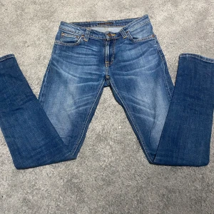 Blå jeans från Nudie Jeans, 29/32 - Snygga blå jeans från Nudie Jeans med klassisk femficksdesign och coola orangea sömmar på bakfickorna. Modellen har en slim fitoch normal midja. Jeansen är tillverkade i slitstarkt denim och har subtila slitningar framtill för en avslappnad vibe. Skick 10/10