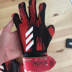 Adidas Predator målvaktshandskar röd/svart - Säljer ett par Adidas Predator målvaktshandskar i rött och svart med vita detaljer. Handskarna har coolt grafiskt mönster och breda kardborreband med Predator-logga. Perfekta för fotbollsmålvakter som vill sticka ut på planen.