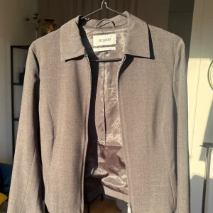 Grå kavajjacka från Weekday - Snygg grå kavajjacka från Weekday med klassisk krage och diskret dragkedja framtill. Jackan har en stilren, rak passform och är helfodrad med ett mjukt innerfoder. Minimalistisk och clean stil.