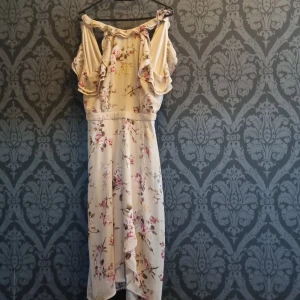 Blommig långklänning från NLY Trend - Säljer en romantisk långklänning från NLY Trend i ljus beige med blommigt mönster i rosa och gult. Klänningen har volangdetaljer vid axlarna, omlott framtill och v-ringad rygg. Perfekt för sommarens alla tillfällen, superfin passform och flowy material.
