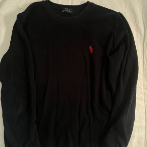 Svart tröja från Polo Ralph Lauren - Svart långärmad tröja från Polo Ralph Lauren med klassisk rund hals och röd broderad logga på bröstet. Tröjan är tillverkad i mjuk bomull och har en clean, stilren look som funkar till jeans eller chinos. Perfekt för dig som gillar enkel och snygg design.