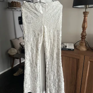 Vita blommiga vida byxor - Supersnygga vita byxor med broderade blommor och lätt glansig finish. Vida ben och hög midja ger en trendig siluett. Perfekta för dig som vill sticka ut med ett unikt plagg. Materialet känns lyxigt och mönstret är riktigt detaljerat.