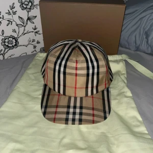 Burberry keps - Säljer en klassisk Burberry keps med ikoniskt rutigt mönster i beige, svart, vitt och rött. Kepsen har justerbar snapback och Burberry London England-logga baktill. Materialet är mjuk bomull och kepsen kommer med original dustbag och box 10/10skick.