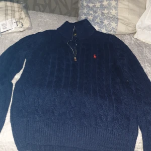 Marinblå stickad tröja Polo Ralph Lauren - Säljer en marinblå stickad tröja från Polo Ralph Lauren med klassiskt kabelstickat mönster och halv dragkedja vid halsen. Tröjan har lång ärm, ribbade muddar och den ikoniska röda Polo-loggan broderad på bröstet. Perfekt för dig som gillar stilren och tidlös design.