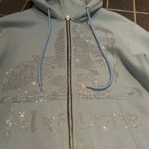 Ljusblå rhinestone hoodie från Maseme - Säljer en ljusblå hoodie från Maseme med glittrande strasstenar på framsidan och coolt motiv. Tröjan har huva med snörning, dragkedja och fickor framtill. Perfekt för dig som gillar att sticka ut med lite bling i vardagen.