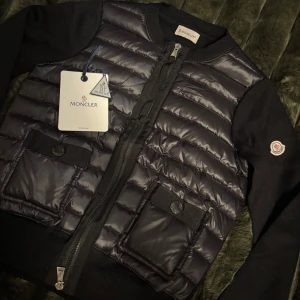 Moncler cardigan - Säljer pga köpte för liten storlek, köpt här på Plick därav kan jag ej returnera. Köpt för 7500kr, när jag fick hem den var den helt oanvänd med plast kvar på dragkedjan längst ner men tog av plasten då jag använt den 2 gånger, därav säljer jag vidare för 1500kr mindre. Den har äkthetsbevis via NFC scan och QR kod, skriv för fler bilder 