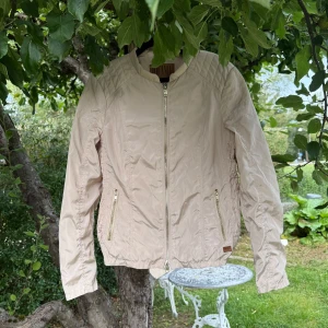 Beige bomberjacka från Jofama - Snygg beige bomberjacka från Jofama med quiltade detaljer på axlarna och dragkedjor i silver både framtill och vid fickorna. Jackan är tillverkad i polyester och har en stilren, sportig look med långärmade ärmar och rund krage. Storlek 42. Mycket fint skick, kan ha små tecken på använd. Men fortfarande fint. Från märket jofama. 