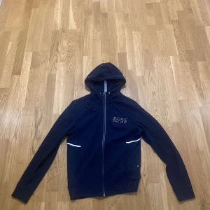 Mörkblå hoodie från Hugo Boss - Snygg mörkblå hoodie från Hugo Boss med vit logga på bröstet och vita detaljer vid fickorna. Tröjan har huva med snörning, hel dragkedja och långa ärmar. Perfekt för chill eller streetwear. Materialet känns mjukt och skönt mot huden.