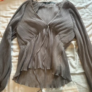 Grå plisserad blus från Zara - Säljer en snygg grå plisserad blus från Zara med v-ringning och långa ärmar. Blusen har knyt framtill och ett flowigt, asymmetriskt fall som ger en cool vibe. Perfekt för dig som gillar detaljer och mjuka material.
