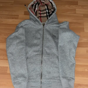 Grå Burberry hoodie med dragkedja - Snygg grå hoodie från Burberry med klassiskt rutigt foder i huvan. Tröjan har hel dragkedja framtill, två fickor och ribbade muddar. Perfekt för dig som gillar stilren design med en touch av lyx. Passar dig som vill ha en chill och exklusiv look.