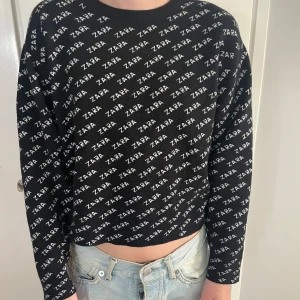 Svart croppad tröja från Zara - Svart croppad tröja från Zara med vit logotyp över hela plagget. Tröjan har lång ärm och rund halsringning, perfekt för dig som gillar streetstyle och vill sticka ut. Materialet är mjukt och skönt, passar till jeans eller kjol.