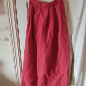 Röd långkjol i ull - Säljer en röd långkjol i ull med knapp och dragkedja på baksidan. Kjolen har en rak passform som samlas i midjan. Den har inget undertyg så är menad att ha med ett par underbyxor eller en underkjol. Den är perfekt att ha de stunder man vill ha kjol under vintern men inte vill frysa.