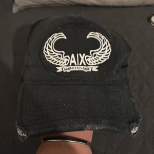 Armani  keps  - Svart keps från Armani Exchange med vit broderad logga och vingar framtill. Har inte använt kepsen på länge, därav säljer jag den. Den är inte trasig eller smutsig utan det är modellen som är så. Hör av er vid frågor! 