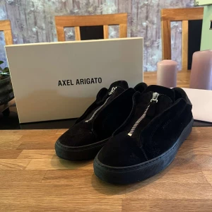 Svarta sneakers Axel Arigato med dragkedja - Snygga svarta sneakers från Axel Arigato med cool dragkedja framtill istället för klassisk snörning. Ovandelen är i mjuk mocka och insidan i skinn, vilket ger en lyxig känsla. Platt sula och rund tå gör dem både stilrena och bekväma.