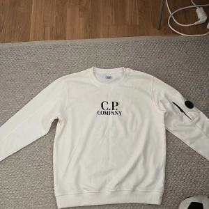 Vit sweatshirt från C.P. Company - Säljer en vit sweatshirt från C.P. Company med svart logga på bröstet och en ikonisk linsdetalj på ärmen. Tröjan har rund halsringning och ribbade muddar. Perfekt för en stilren och modern look. Använd bara en gång och är i jättebra skick.
