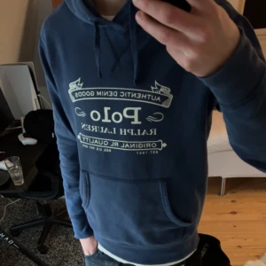Blå hoodie från Polo Ralph Lauren - Säljer en snygg marinblå hoodie från Polo Ralph Lauren med vit tryckt logga på bröstet. Tröjan har huva med snörning och en stor magficka framtill. Perfekt för dig som gillar streetwear och vill ha en klassisk look.