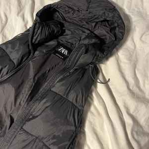 Svart pufferväst från Zara - Säljer en svart pufferväst från Zara med huva och dragkedja. Jackan har quiltad design, praktiska fickor med dragkedja och en loose passform. Snygg och enkel att matcha med din vardagsstil.