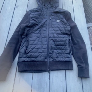 Svart quiltad pufferjacka J.Lindeberg (Cardigan) - Svart pufferjacka från J.Lindeberg med quiltad framsida, huva och dragkedja. Jackan har ribbade muddar, två sidofickor med dragkedja och en snygg logga på bröstet. Materialet är polyester och jackan har en clean, modern look som passar till streetwear.