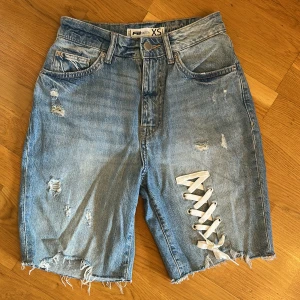 Blå jeansshorts med snörning  - Snygga blå jeansshorts i storlek XS. Modellen är med råa kanter och slitna detaljer. På ena benet finns en vit snörning som ger en cool touch;)