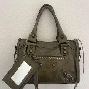 Olivgrön handväska  - Snygg olivgrön handväska i äkta skinn, tillverkad i Italien. Väskan har coola metalldetaljer, dragkedja framtill och ett praktiskt fack med spegel. Väskan är i nyskick och köptes för 900kr