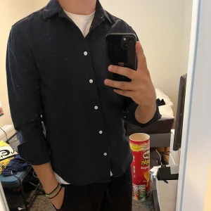 Mörkblå bomullsskjorta från H&M - Snygg mörkblå skjorta från H&M i mjuk bomull. Klassisk krage, vita knappar och långärmad modell med regular fit. Perfekt att bära över en t-shirt för en chill och stilren look.