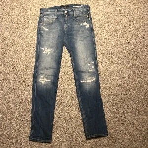 Replay Anbass blå slim jeans - Snygga blå jeans från Replay, modell Anbass med slim fit. De har slitna detaljer och snyggt tvättad look, klassiska fem fickor och Replay-logga på bakfickan. Perfekt passform med smal siluett och coola slitningar på benen.