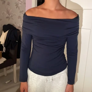 Mörkblå offshoulder långärmad topp - Snygg mörkblå offshoulder topp med långa ärmar och draperad halsringning. Toppens tighta passform framhäver silhuetten och den är gjord i ett mjukt, stretchigt material. Perfekt för dig som gillar stilrena och trendiga plagg. Storlek 146/152 men passar xs-s. 