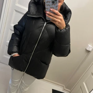 Svart pufferjacka från Massimo Dutti - Säljer en svart pufferjacka från Massimo Dutti med stor huva och coolt snedställd dragkedja. Jackan är quiltad och har en oversized vibe, perfekt för kalla dagar. Materialet är glansigt och känns riktigt mjukt och skönt.