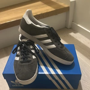 Adidas Gazelle grå sneakers - Säljer ett par Adidas Gazelle sneakers i mörkgrå mocka med vita detaljer och klassiska tre ränder på sidan. Skorna har mörkgrå snörning kommer även med i vit, vit platt sula och rund tå. De är helt nya använda en gång säljer på grund av att de va för stora kontakta vid fler frågor eller bilder