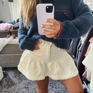 Vit volangkjol med fickor - Supersöt vit kortkjol med volangkant och praktiska fickor fram. Kjolen har en knapp och dragkedja i midjan och är gjord i ett mjukt bomullstyg som känns skönt mot huden. Perfekt för dig som gillar en chill och feminin stil.