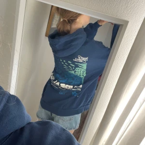 Blå hoodie med tryck  - Säljer en mörkblå hoodie med coolt tryck i vitt och turkos på både fram- och baksidan. Tröjan har huva, ribbade muddar och en relaxed, loose passform. Perfekt för chill dagar och streetstyle.
