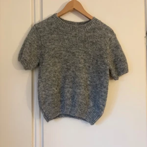 Grå stickad tröja från Gina Tricot - Mysig grå stickad tröja från Gina Tricot i storlek S. Tröjan har korta ärmar, rund hals och ribbade muddar vid ärmslut och nederkant. Perfekt för lager-på-lager och chill dagar. Materialet är mjukt och fluffigt, vilket gör den extra skön att bära.