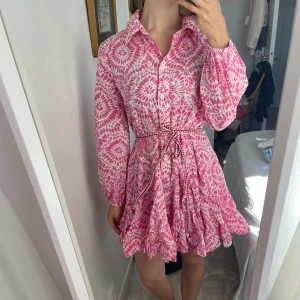 Rosa och vit kortklänning med snörning - Säljer en superfin kortklänning i rosa och vitt med blommigt mönster. Klänningen har långärmade ärmar, krage och knappar framtill. Midjan markeras med en snörning och kjolen har volangdetaljer som ger en snygg och lekfull look.